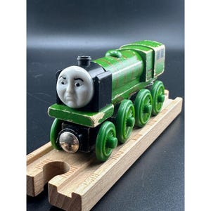 May include: A green wooden toy train with a white face and black accents. The train has green wheels and is sitting on a wooden track. The train is a classic toy, perfect for children.
