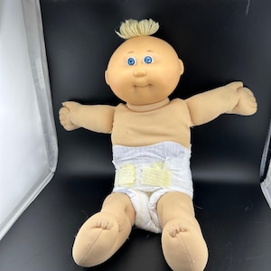 Op de afbeelding: Een pop met een zacht lichaam, een rond, perzikkleurig gezicht, helderblauwe ogen en een klein plukje blond garen op het hoofd. De pop heeft uitgestrekte armen en draagt een witte wegwerpluier met gele plakstrips.