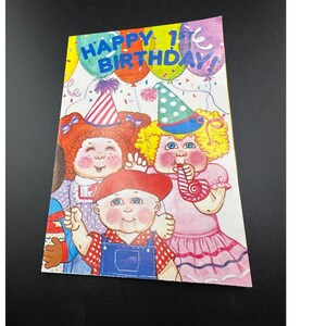 May include: A vintage birthday card featuring three Cabbage Patch Kids. The card has the text "HAPPY 1st BIRTHDAY!" above the characters. The characters are wearing party hats and holding party favors. The background includes colorful balloons.