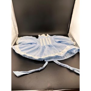 Peut inclure: Une robe de poupée bleu clair avec des manches courtes bouffantes, un col montant et une bordure en dentelle blanche à œillets le long de l'ourlet et du corsage. Le vêtement présente des détails en dentelle verticale et deux longs liens.