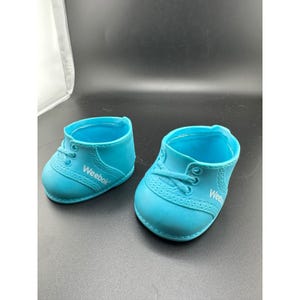 May include: A pair of bright cyan blue molded plastic doll shoes featuring a low-top sneaker design with molded laces and eyelets. The side of each shoe displays the white text Weebok in a clean, sans-serif font, mimicking a classic athletic style.