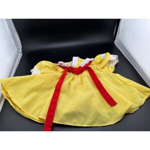 May include: A vintage yellow dress with a red bow and red ribbons. The dress has white lace trim around the collar and sleeves. The dress is made of a lightweight fabric.