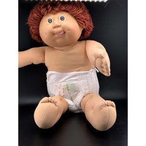 May include: A vintage Cabbage Patch Kid doll with brown hair, a smiling face, and a diaper. The doll is sitting with its legs extended. The doll's skin is a light tan color.