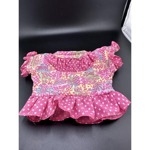 Op de afbeelding: Een top met korte mouwen voorzien van een levendig, abstract geometrisch patroon in roze, geel en blauw. Het kledingstuk heeft een ruchekraag en een brede, uitlopende zoom met een witte stippenprint op een roze achtergrond.
