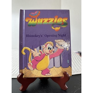 Könnte beinhalten: Ein Vintage-Buch "Wuzzles" mit dem Titel "Rhinokey's Opening Night" auf einem Holzständer. Das Cover ist lila mit einer Cartoon-Illustration eines rosa und gelben Charakters. Das Buch gehört zur "Collector Series".