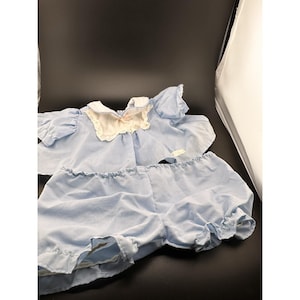 Pode incluir: Um conjunto de boneca azul claro, com uma parte superior com gola com acabamento em renda branca e calções a condizer. A parte superior tem mangas curtas e um detalhe de laço. Os calções têm uma bainha com folhos. O conjunto é feito de tecido macio e leve.