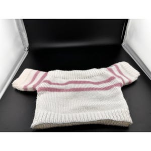 May include: A small, off-white knitted sweater with horizontal stripes in a muted pink color. The short-sleeved top is made of a soft, textured yarn, perfect for a doll or small toy. The sweater has a simple, classic design.