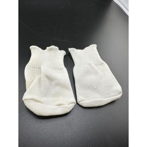 May include: A pair of white infant mittens made from a soft knit fabric. Each mitten features a ribbed elastic cuff at the wrist to ensure a secure fit, with a rounded, seamless design that covers the entire hand area for protection.