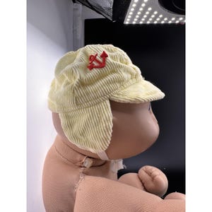 May include: A light yellow corduroy cap with a red emblem. The cap has ear flaps and a small peak. The cap is on a doll.