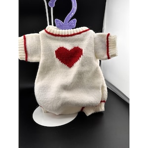 May include: A white knit romper with a large red heart on the front. The romper has red trim around the neckline, sleeves, and leg openings. The garment is displayed on a purple hanger.