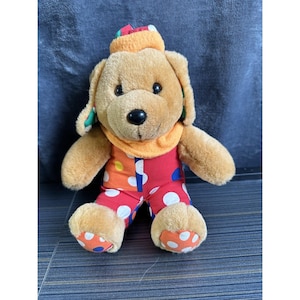 May include: A plush dog toy with a brown body, wearing a colorful outfit. The dog has a round black nose, floppy ears, and a cheerful expression. It is dressed in a red jumpsuit with white polka dots, an orange collar, and a matching hat.