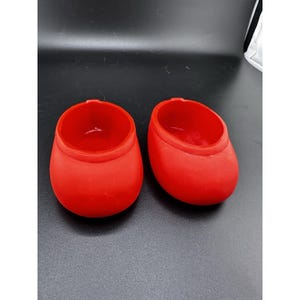 Può includere: Due piccoli contenitori ovali di colore rosso vivo. Hanno basi arrotondate e ampie aperture. I contenitori, realizzati in un materiale liscio, poggiano su una superficie scura. La loro forma ricorda delle piccole scarpe. Circa 5 cm di altezza.