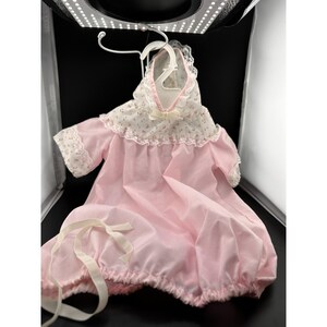 Op de afbeelding: Een vintage roze babyjurk met een bloemenpatroon op de capuchon en mouwen, afgezet met kant. De jurk heeft een witte strik bij de halslijn en een gerimpelde rok. Het hangt aan een witte hanger.