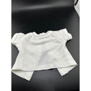 Pode incluir: Uma camisa branca de manga curta com decote redondo e costuras decorativas cinza-escuras ao longo da gola e das bordas das mangas. A peça tem um corte simples e reto, projetada para uma boneca pequena ou figura semelhante.