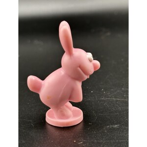 May include: A pink plastic bunny figurine with a smiling face, white eyes, and a red nose. The bunny is standing on a round base. The figurine has a cartoonish design.