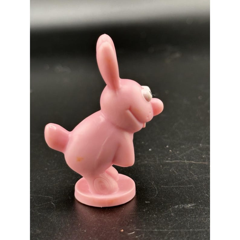 May include: A pink plastic bunny figurine with a smiling face, white eyes, and a red nose. The bunny is standing on a round base. The figurine has a cartoonish design.
