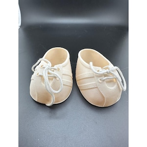Peut inclure: Une paire de baskets pour poupée de couleur crème avec une surface texturée, des bouts arrondis et des lacets blancs passés dans des œillets. Les chaussures présentent un design bas avec des bandes latérales moulées et une structure solide.