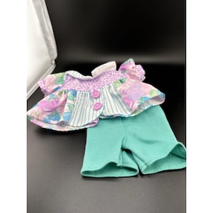 May include: A doll's outfit featuring a floral print top with purple buttons and a turquoise shorts. The top has a white collar and a mix of floral and striped patterns. The shorts are a solid turquoise color.
