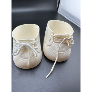 May include: A pair of off-white, moulded plastic doll shoes featuring a rounded toe, a decorative embossed heart pattern on the vamp, and white fabric laces threaded through eyelets. The shoes have a high-top silhouette with an open top.
