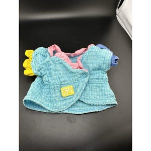 Könnte beinhalten: Hellblaues Baby-Doll-Oberteil aus strukturiertem Stoff. Das Oberteil hat rosa und gelbe Akzente am Ausschnitt und an den Ärmeln. Ein kleines gelb-blaues Logo ist auf der Vorderseite zu sehen.