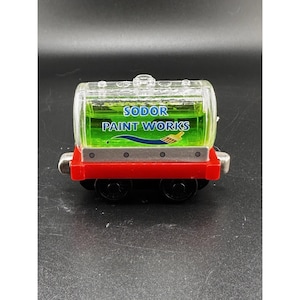 May include: A toy train car from the Sodor Paint Works. The car has a red base with black wheels and a clear, rounded top filled with green liquid. The text "SODOR PAINT WORKS" is printed on the top.