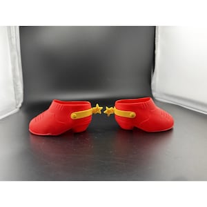May include: A pair of red toy cowboy boots with yellow spur attachments. The boots have a rounded toe and a small heel. The spurs are star-shaped and attached with a yellow band.