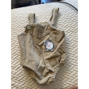 Op de afbeelding: Een beige corduroy romper met een mouwloos ontwerp, twee schouderbandjes en een enkele witte knoopsluiting aan de voorzijde. Een geborduurde patch van een ronde olifant met een roze oor en een grijs lichaam bevindt zich aan de rechteronderkant.