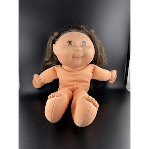 Peut inclure: Une poupée Cabbage Patch Kid vintage avec des cheveux bruns et un corps de couleur pêche. La poupée a une expression souriante et est assise, les jambes tendues. Les yeux de la poupée sont bruns et elle a un petit visage rond.