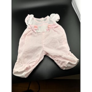 Könnte beinhalten: Ein rosa-weiß gestreifter Baby-Strampler mit kurzen Ärmeln und Rüschenbündchen. Das Oberteil ist weiß mit einer dekorativen rosa Schleife und gestickten Details. Das Kleidungsstück ist für Säuglinge konzipiert.