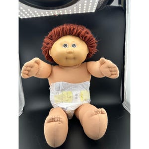 May include: A vintage Cabbage Patch Kid doll with a tan complexion, brown hair, and blue eyes. The doll is wearing a diaper with yellow accents. The doll is sitting with its arms and legs outstretched.