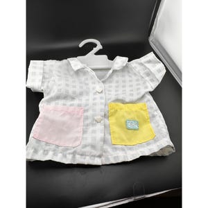 May include: A doll-sized, short-sleeved shirt with a white and gray checkered pattern. It features a pink pocket on the left and a yellow pocket on the right. The shirt is buttoned and hangs on a white plastic hanger.