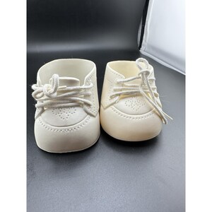 May include: A pair of off-white doll shoes featuring a rounded toe, a perforated heart-shaped design on the vamp, and white fabric laces tied in a bow. The shoes have a smooth, moulded construction with visible stitching details along the edges.