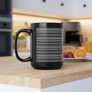 Könnte beinhalten: Schwarze Keramiktasse mit weißem geometrischen Muster. Das Design zeigt horizontale Linien unterschiedlicher Breite, wodurch ein strukturiertes Aussehen entsteht. Die Tasse hat einen großen Henkel und steht auf einer Holzoberfläche.