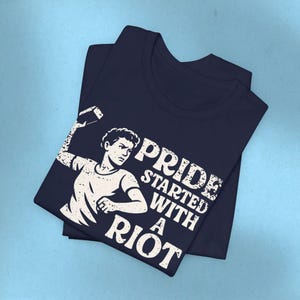 May include: Navy blue t-shirt with a distressed white graphic of a person throwing a brick, and the text "PRIDE STARTED WITH A RIOT". The shirt is folded and stacked on a light blue surface.