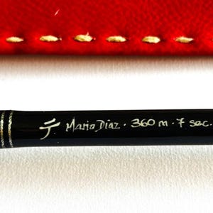 Op de afbeelding: Zwarte hengel met zilveren details en de tekst "Mario Diaz · 360 m · 7 sec." De hengel staat tegen een rode achtergrond met witte stiksels.