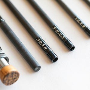 Op de afbeelding: Close-up van verschillende zwarte hengelstoksecties met witte tekst. De hengels zijn schuin, met de uiteinden naar rechts gericht. De handgreep van één hengel is links zichtbaar, met een kurken handvat. De tekst op de hengels luidt "8'3 #5".