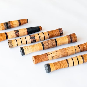 Puede incluir: Seis tapones de corcho para botellas con varios diseños. Los tapones son cilíndricos, con corcho natural y detalles oscuros. Algunos tienen extremos negros o estampados, mientras que otros tienen un acabado de corcho liso. Están dispuestos en diagonal sobre una superficie blanca.