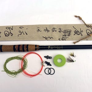 Puede incluir: Una caña de pescar negra con mango de corcho, una funda de tela beige con caligrafía negra, un carrete de hilo verde, dos clips negros, dos anillos negros, dos moscas de pesca y sedales de pesca enrollados en verde y rojo.