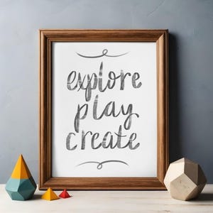 May include: A framed print with the words "explore play create" in a handwritten style. The frame is brown, and the print is on a white background. Decorative wooden geometric shapes in yellow, red, and blue are in front of the frame.
