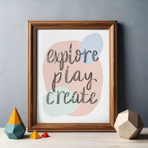 May include: A framed print with the words "explore play create" in a handwritten style. The background has pastel shapes in pink, blue, and green. Wooden geometric shapes are on the table.