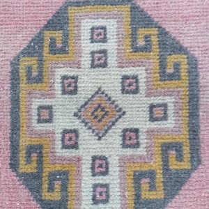 Puede incluir: Una alfombra geométrica rosa y gris con un diseño octogonal central. El patrón presenta una forma de cruz con cuadrados y un diamante en el centro, delineado en amarillo y gris.