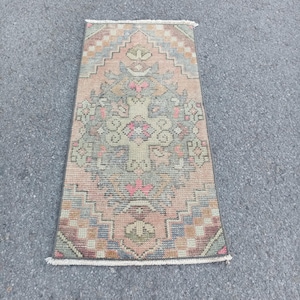 May include: A rectangular woven rug featuring a central diamond medallion pattern in muted shades of peach, sage green, dusty blue, and cream. The rug has a symmetrical geometric design with a fringed edge on the top and bottom short sides.