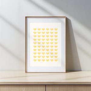 May include: A framed wall art print featuring a grid pattern of small, light yellow hearts arranged in ten rows and five columns on a cream-colored background, enclosed within a simple, thin, light-toned wooden frame.