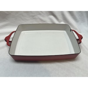 May include: A rectangular baking dish with a vibrant red exterior and a smooth, off-white interior. The dish features two integrated red handles on opposite sides and a dark rim along the top edge, providing a clean and functional design.