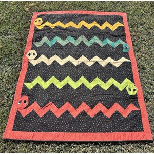 May include: A rectangular quilt featuring a black background with small multicolored dots. Four horizontal zigzag-shaped snakes in yellow, teal, beige, and lime green are appliqued across the center, with a red zigzag snake at the bottom.