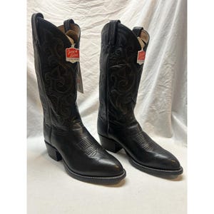 May include: A pair of black leather cowboy boots featuring intricate tonal stitching on the tall shafts, pointed toes, and stacked heels. Each boot includes a red and white Tony Lama brand tag attached to the side, showcasing classic western styling.