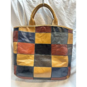 May include: A leather tote bag featuring a patchwork design composed of square panels in various colors including mustard yellow, navy blue, deep red, black, and dark brown. The bag has two matching leather handles attached to the top edge.