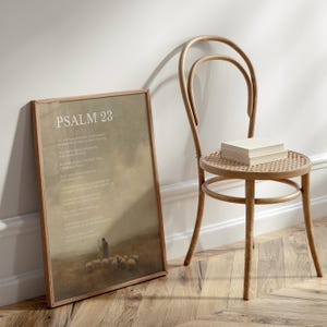May include: A framed print with the text "PSALM 23" in a light-colored font. The print depicts a shepherd with sheep in a field. A wooden chair with a woven seat and a stack of books sits next to the print.