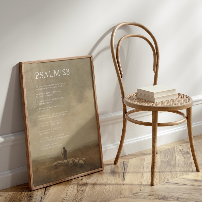 May include: A framed print with the text "PSALM 23" in a light-colored font. The print depicts a shepherd with sheep in a field. A wooden chair with a woven seat and a stack of books sits next to the print.
