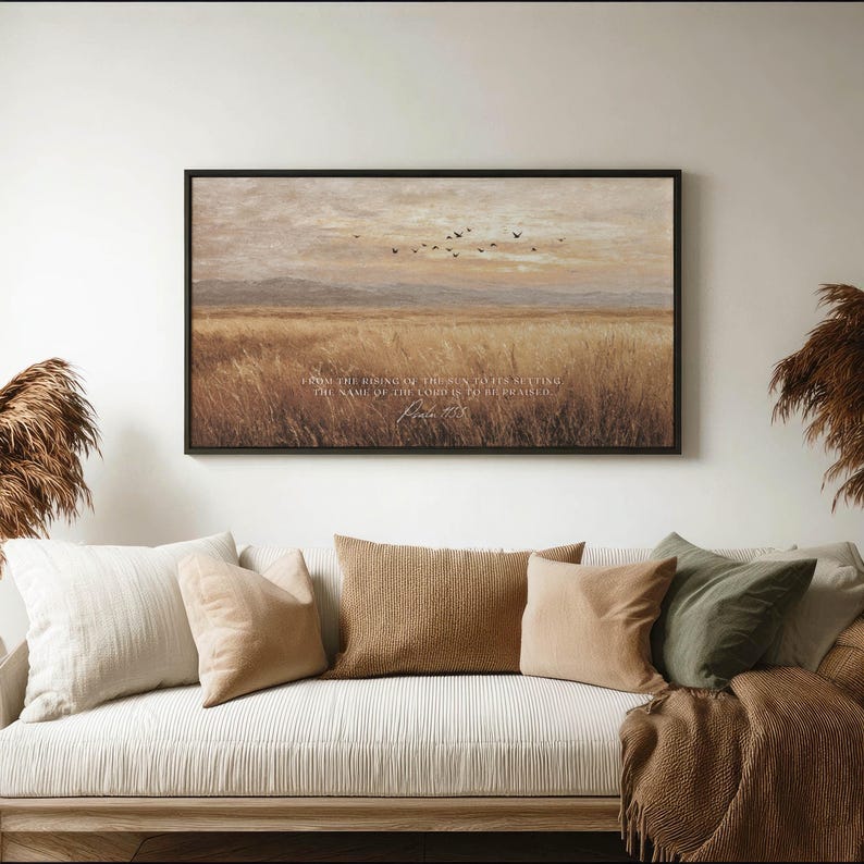 May include: A framed landscape art print featuring a field of tall grass under a muted sky with flying birds. The artwork has a black frame and includes text from Psalm 113:3. The print hangs above a sofa with various throw pillows.